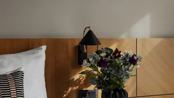 Et koselig hotellrom med seng, sengegavl i tre, nattbord med blomster og abstrakt kunst på veggen, badet i sollys.