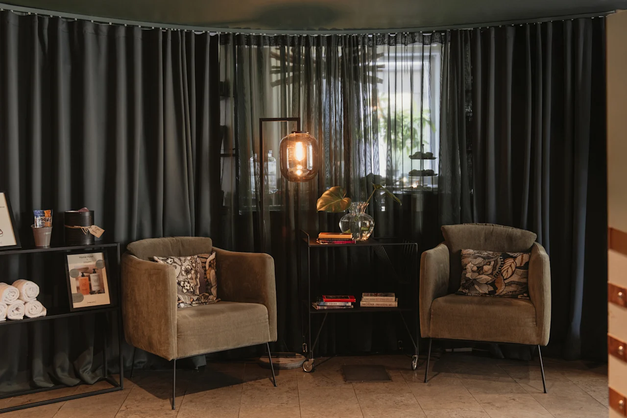 A cozy seating area at Home Hotel Uppsala with two armchairs, a small table, and a warm lamp, set against dark curtains.