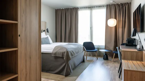 Et pænt møbleret hotelværelse indeholder en seng med gråt sengetøj, et træskrivebord med lampe og kaffemaskine og to stole ved et rundt bord nær et stort vindue med gardiner.