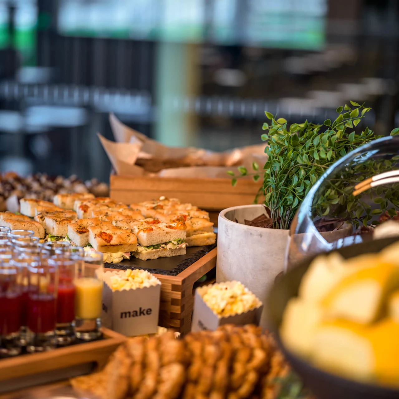 Et buffébord viser assorterte smørbrød, miniburgere og drikker i glass, arrangert på et trebord med dekorative planter. En liten eske med ordet "make" er synlig.