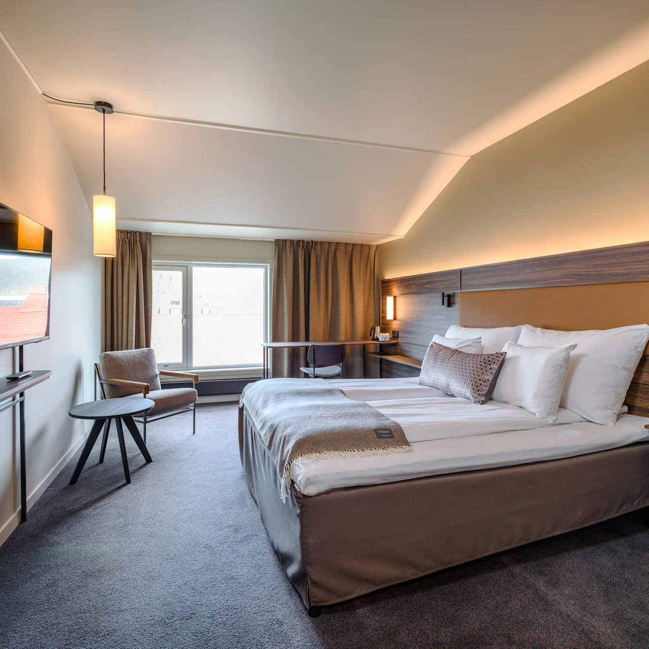 A neatly-made hotel bed with white linens is positioned near a large window, lit by soft lighting. A wall-mounted TV and a small table with a chair complete the modern room.
