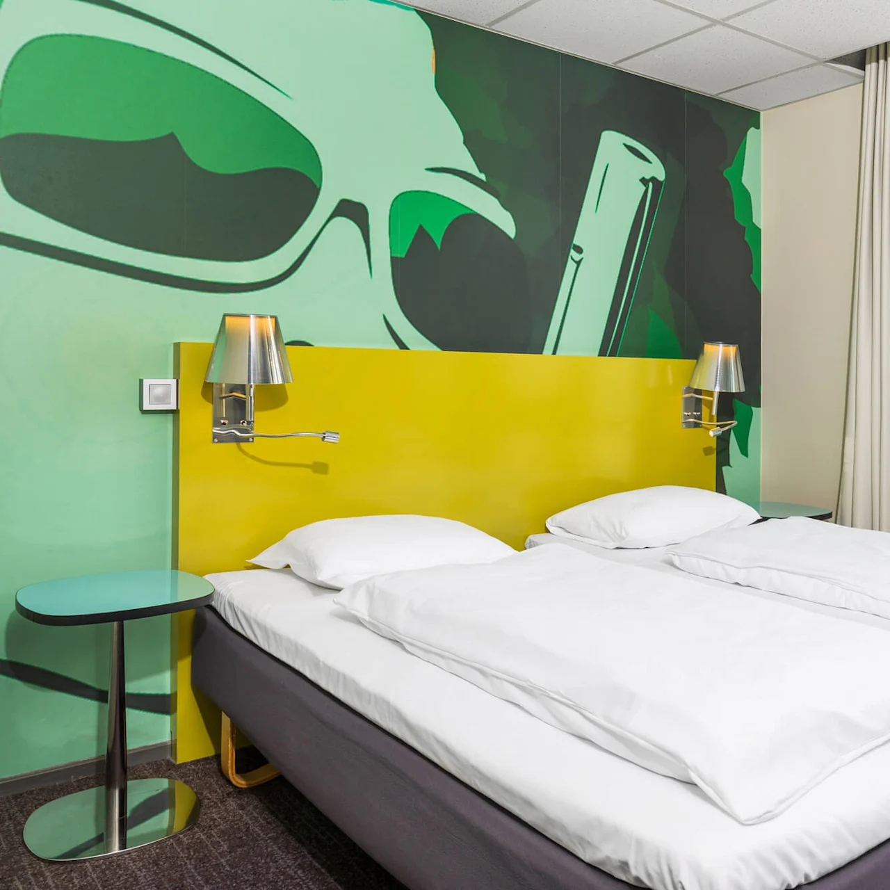 Two white twin beds stand beside a yellow headboard with attached lamps, against a vibrant wall mural depicting sunglasses, in a bright, modern room with a large window.