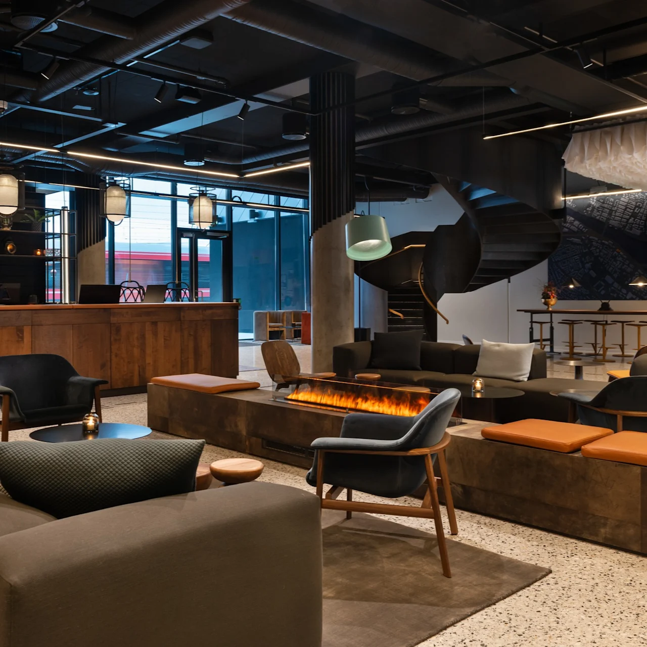 Et moderne lobbyområde med stilfulle sitteplasser rundt en elegant, rektangulær peis. Overhead henger industrielle lys fra taket. En tre-resepsjon og en spiraltrapp er synlige.