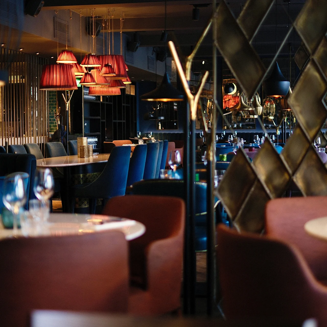 Elegant restaurant tables set with glasses and plates, surrounded by colorful chairs. Warm lighting from hanging lamps creates a cozy atmosphere, with geometric decor in the background.