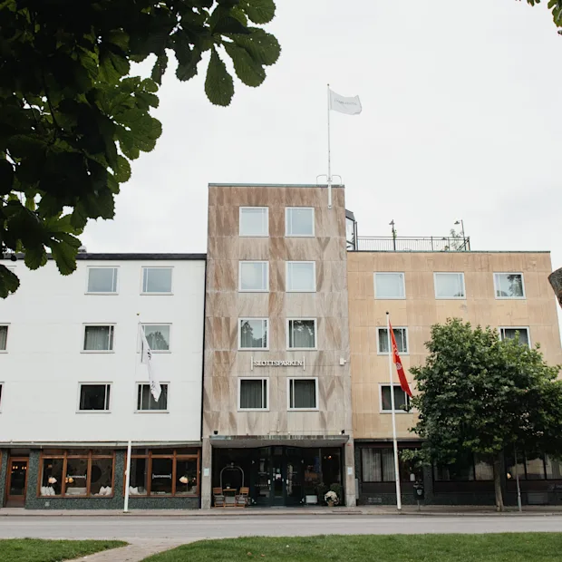 A large building with a sign reading SLOTTSSPARKEN, featuring different facades and flags, framed by green trees.