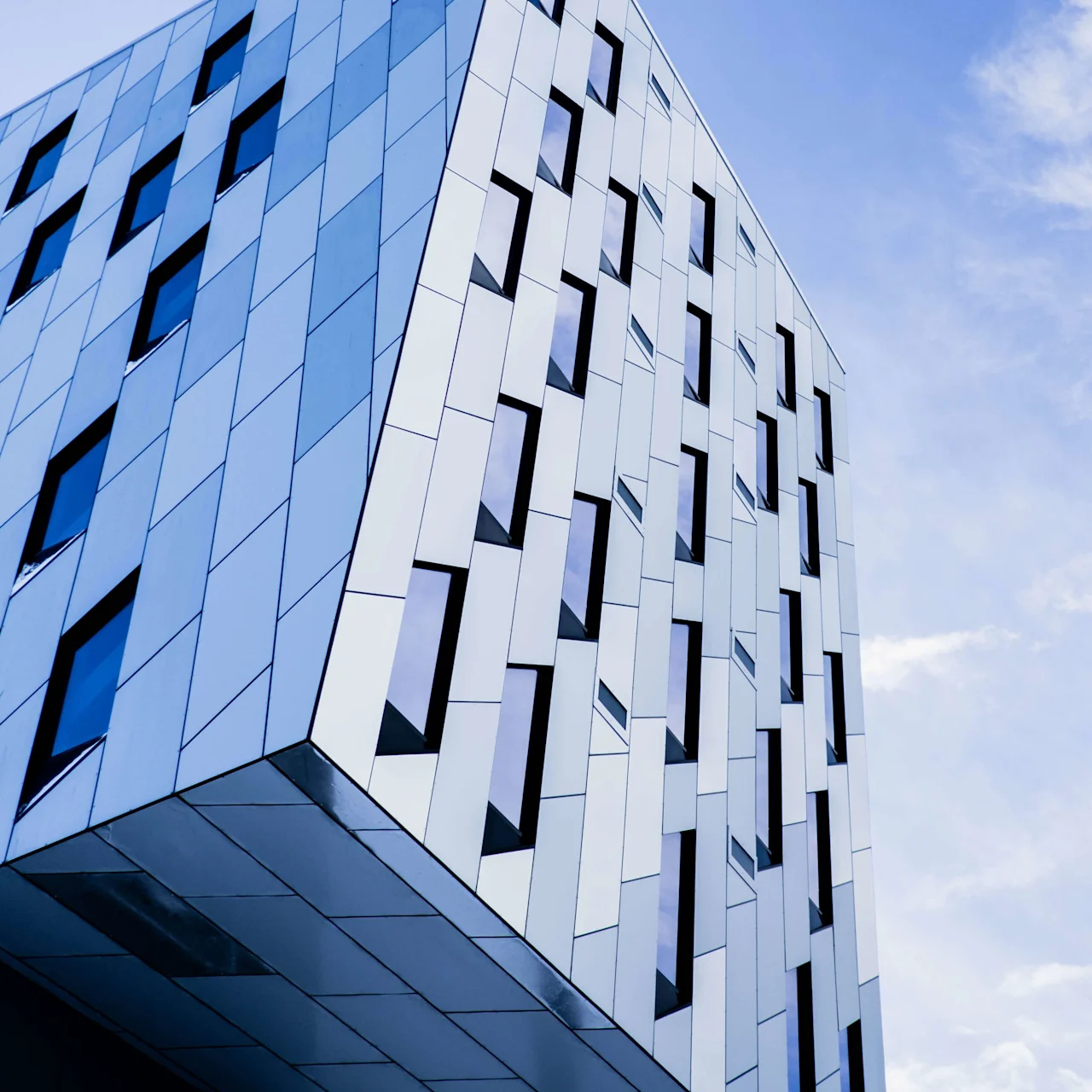 En modern byggnad med geometrisk fasad med alternerande mönster av glas- och metallpaneler, som stiger mot en klarblå himmel.