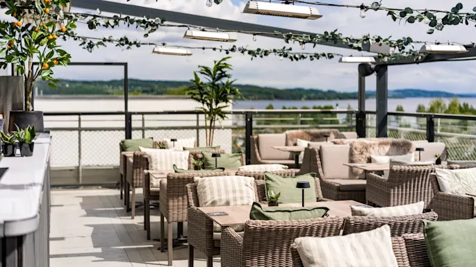 Wicker chairs with cushions line a rooftop terrace, under a pergola adorned with lights and greenery. The backdrop features a scenic view of distant trees and water.