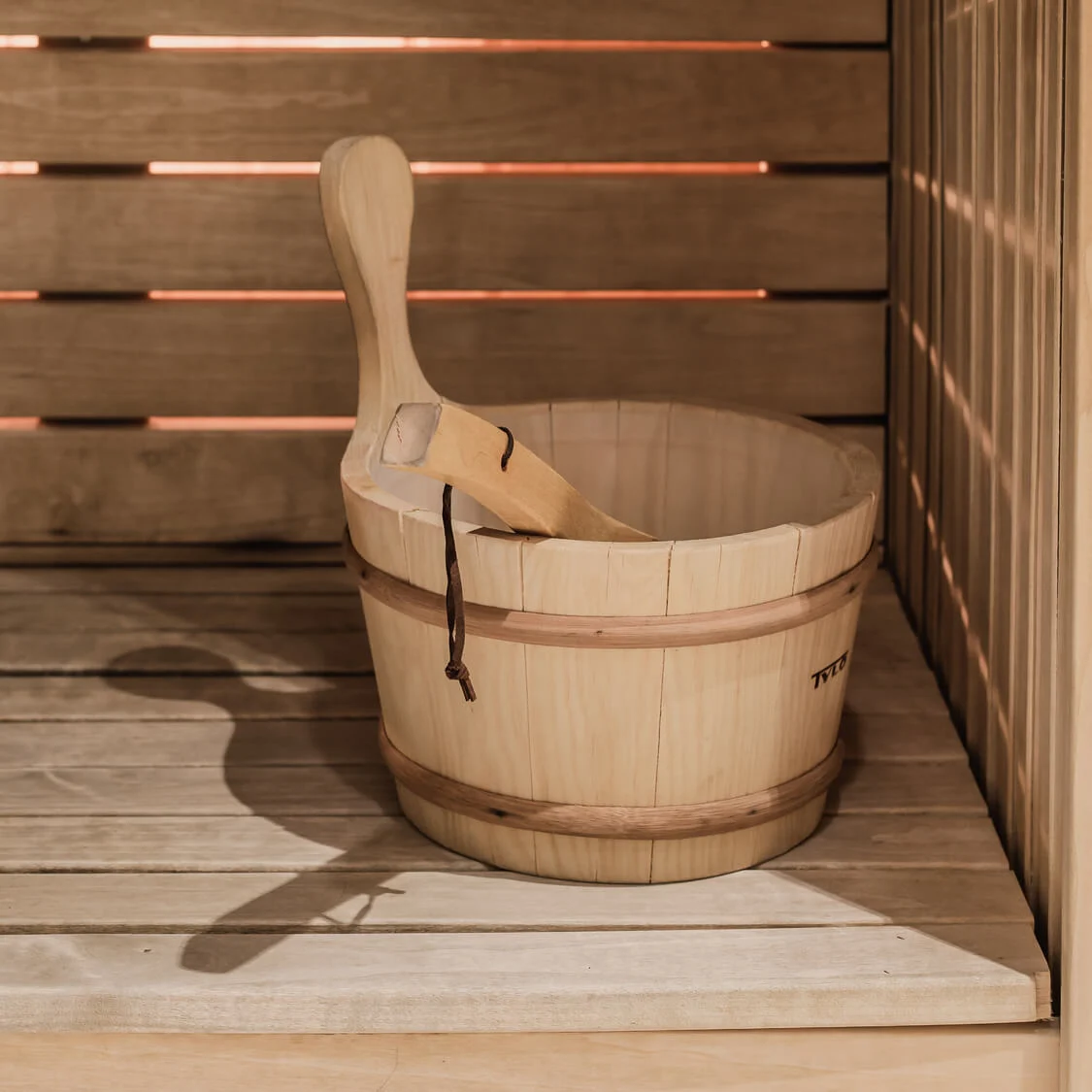 A wooden bucket with a ladle rests on a wooden bench inside a sauna, surrounded by slatted wooden walls.