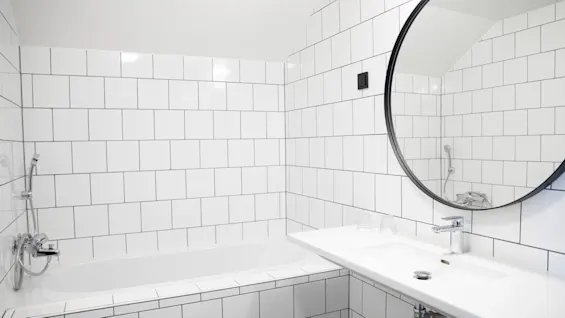 Et hvidt badekar står under en flisebelagt væg, akkompagneret af en vask med et rundt spejl over, i en moderne, minimalistisk badeværelsesindstilling.