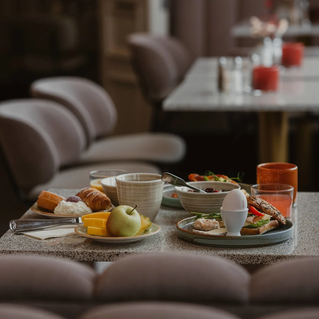 Breakfast foods are arranged on a table, including a croissant, cheese, apple, melon slices, egg, and coffee cup, in a softly lit dining area with cushioned chairs.