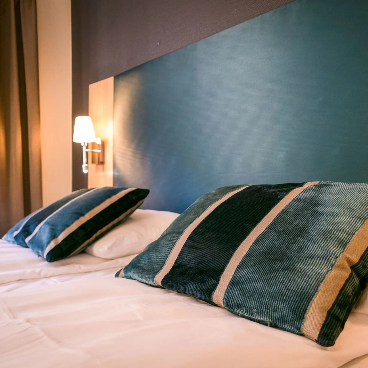 Two striped pillows rest on a neatly made bed with white sheets, set against a teal wall. A lamp illuminates the area, with brown curtains in the background.