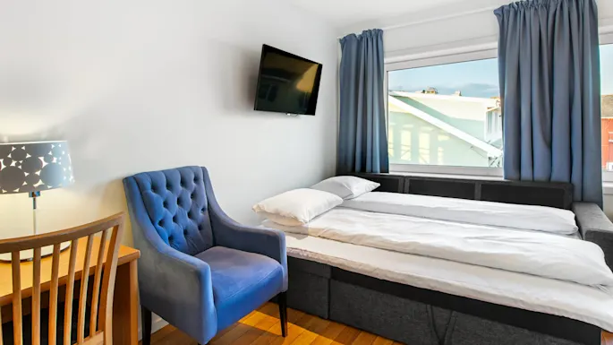 Bed sits beside blue armchair and wooden desk with lamp, facing wall-mounted TV. Blue curtains frame large window overlooking rooftops under clear sky.