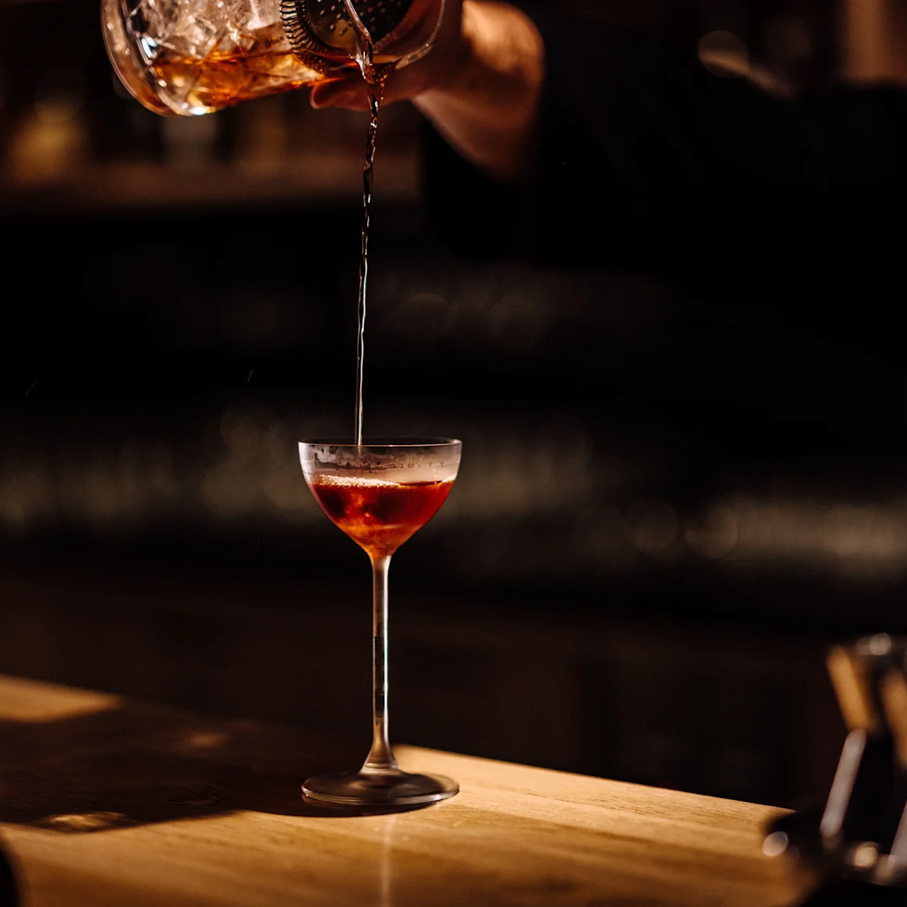 Et cocktailglass får en drink hellet fra en shaker holdt av en hånd, satt på en trebar i et svakt opplyst miljø.