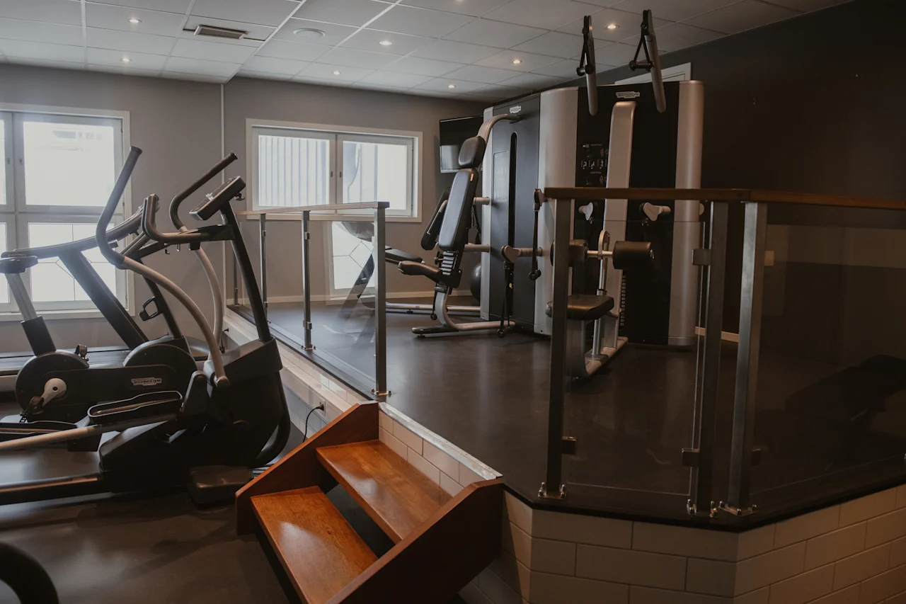 Moderni kuntosali, jossa on erilaisia kuntoilulaitteita, kuten juoksumatto, crosstrainer ja painoharjoittelulaitteet, valoisassa huoneessa, jossa on suuret ikkunat ja jaettu tasorakenne.