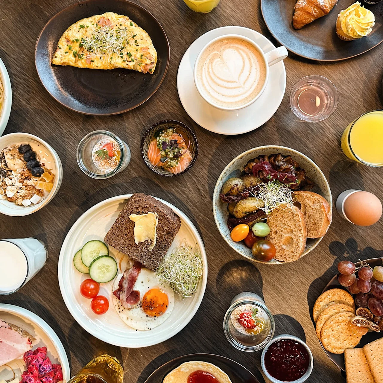 A variety of breakfast foods, including omelettes, fruit, pastries, cheese, and coffee cups, are arranged on a wooden table, suggesting a buffet or brunch setting.