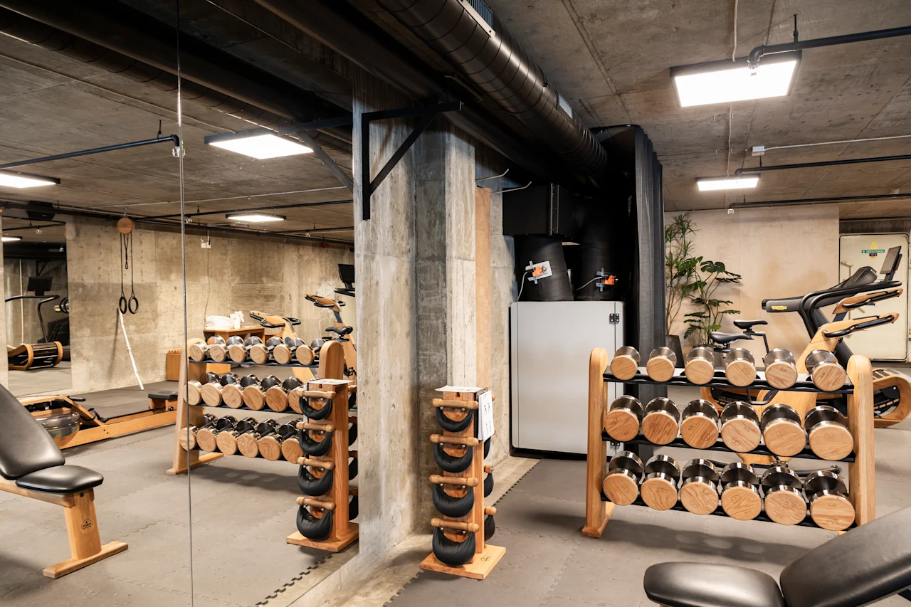 Hantlar och kettlebells står på trärack i ett modernt, rymligt gym med betongväggar och tak. Motionscyklar och rep syns i bakgrunden.