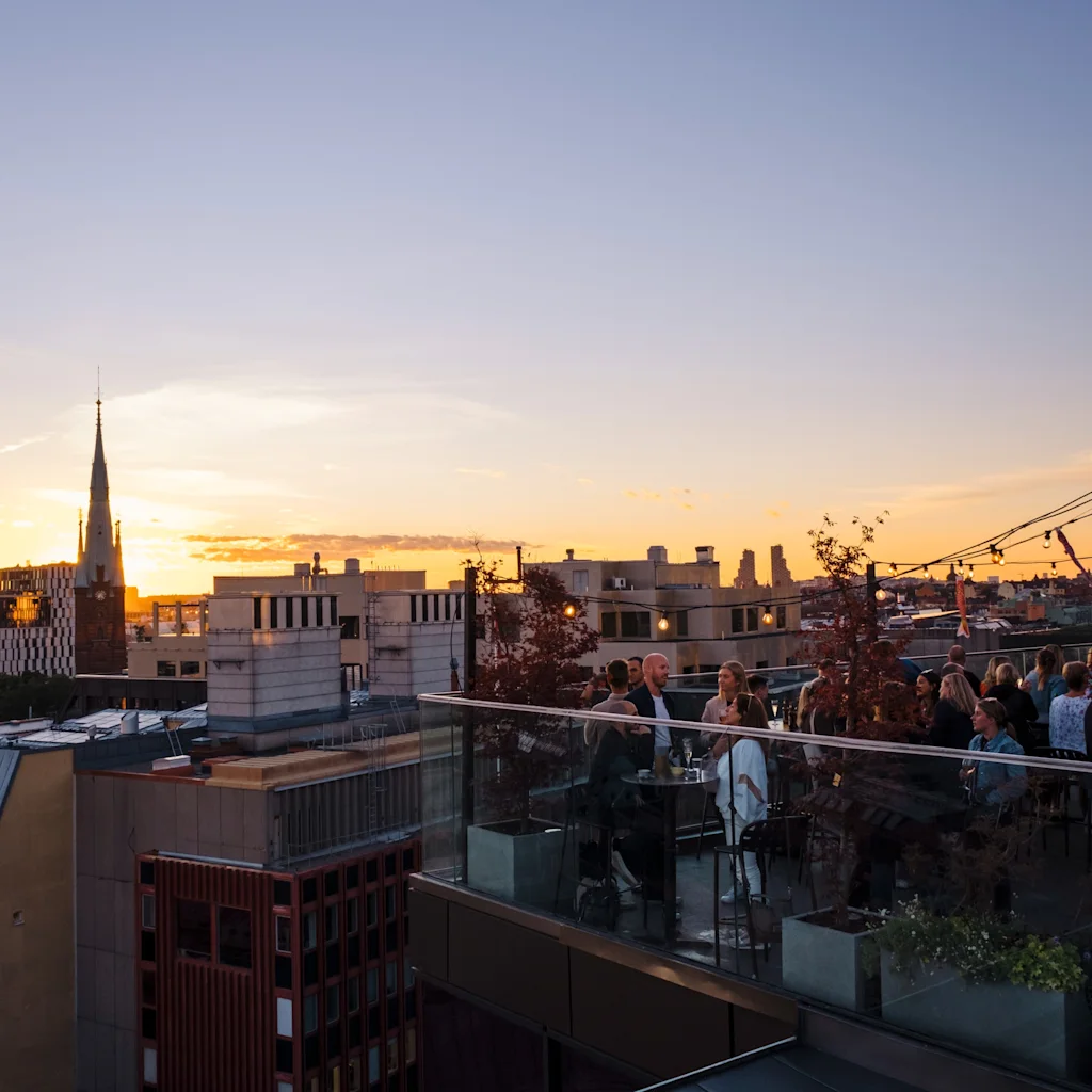 Folk nyter drinker og samtaler på en takterrasse ved solnedgang, med byens silhuett i bakgrunnen.