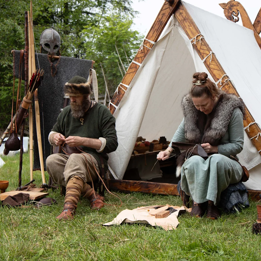 Kaksi historiallisissa asuissa olevaa henkilöä valmistaa huolellisesti nahkatavaroita ulkona, perinteinen teltta taustalla.