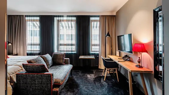 Et moderne hotellrom har en mønstret sofa vendt mot et vindu, en trepult med en rød lampe og stol, og en veggmontert TV.