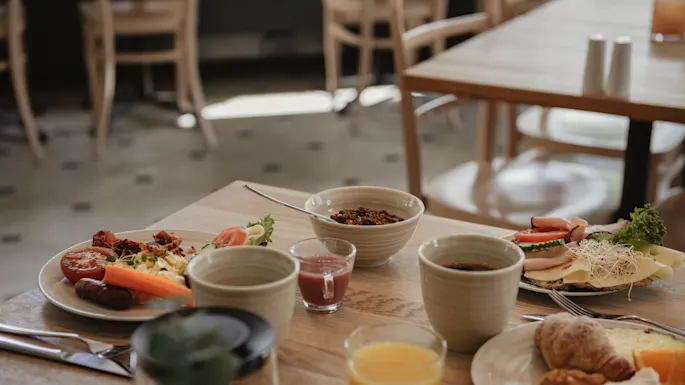 Frokostbord med tallerkener med mat og kaffekopper på et trebord; tomme stoler kan ses i bakgrunnen, noe som skaper en koselig kafeatmosfære.
