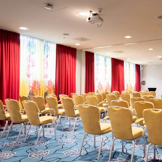 Rader med gule stoler er arrangert i et konferanserom med røde gardiner og blomstrete vinduer, under taklamper og en projektor, på et blått teppe med blomsterdesign.