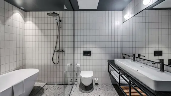 Et moderne badeværelse har et fritstående badekar, en bruser med rundt brusehoved, vægmonteret toilet og håndvask. Rummet har gråt flisebelagt gulv og vægge.