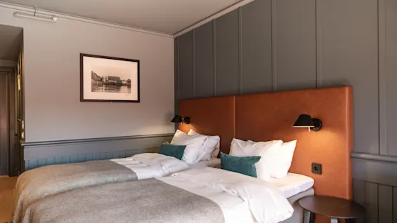 To pænt rederede enkeltsenge med hvidt og gråt sengetøj er placeret under en brun polstret sengegavl. Subtile væglamper oplyser det hyggelige rum, udsmykket med elegante mørke vægge og et indrammet billede.