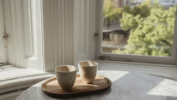 Två keramikkoppar står på en träbricka på ett marmorbord bredvid ett fönster. Solljus filtreras igenom och lyser upp en suddig vy av gröna träd utanför.