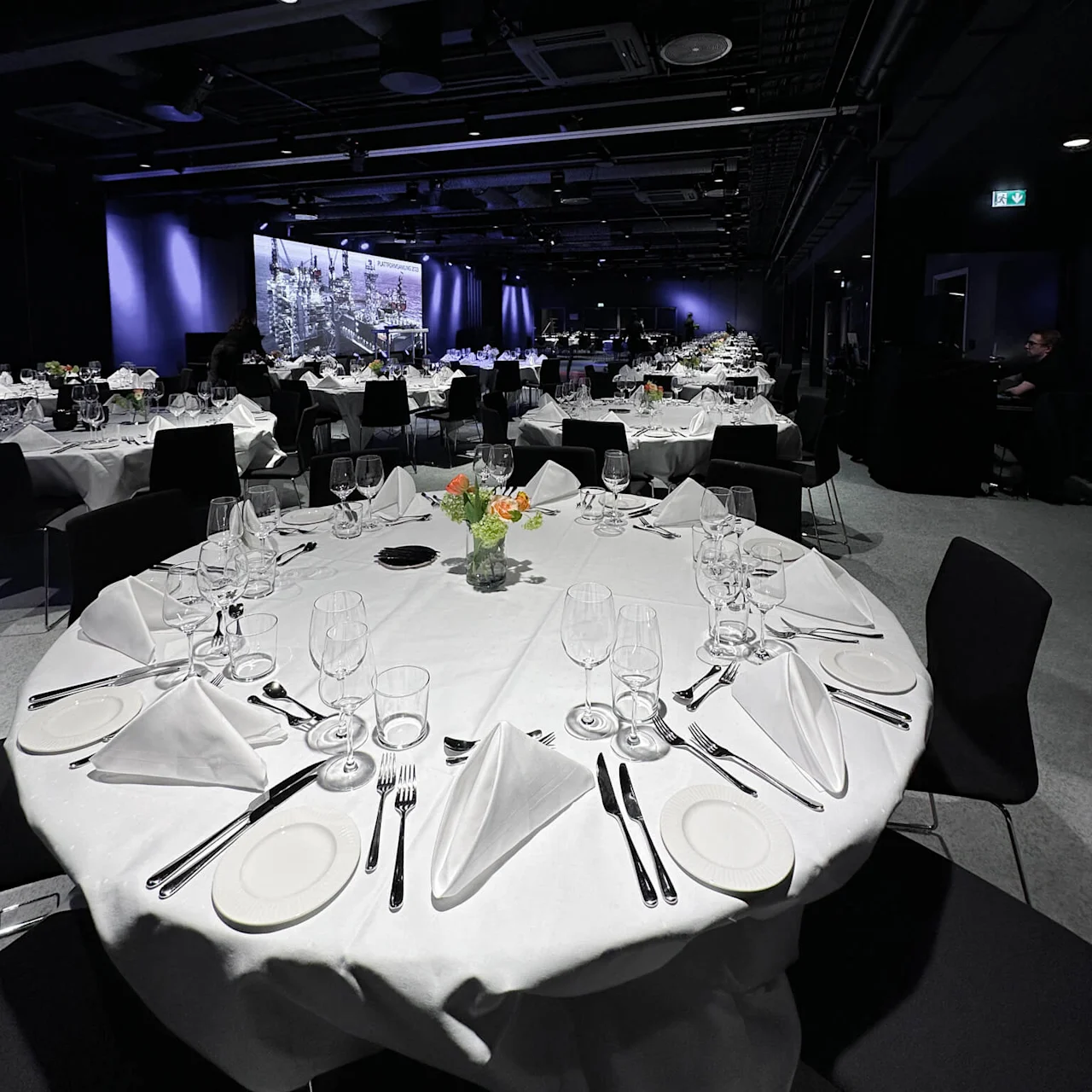 Round tables are elegantly set with white tablecloths, glassware, and folded napkins. Vases with small floral arrangements adorn each table. The dimly-lit hall features a large projection screen.