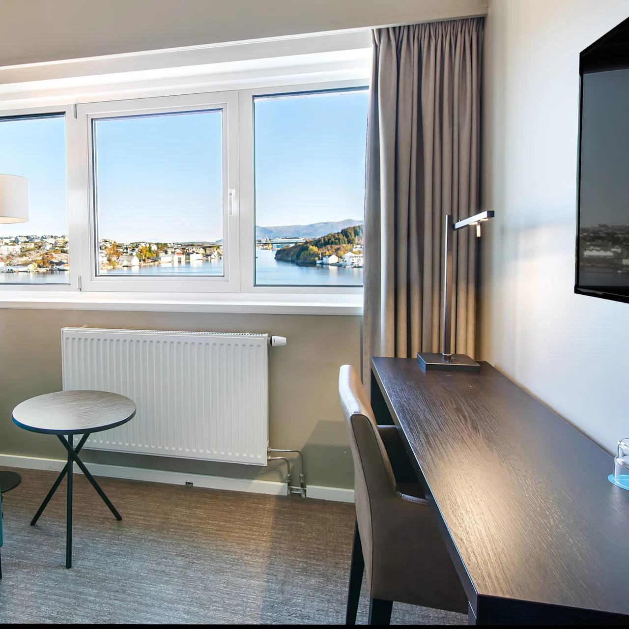 En blå lenestol står ved siden av et lite rundt bord og gulvlampe. Et vindu viser en vakker kystutsikt, og et skrivebord med en lampe og TV er i nærheten.