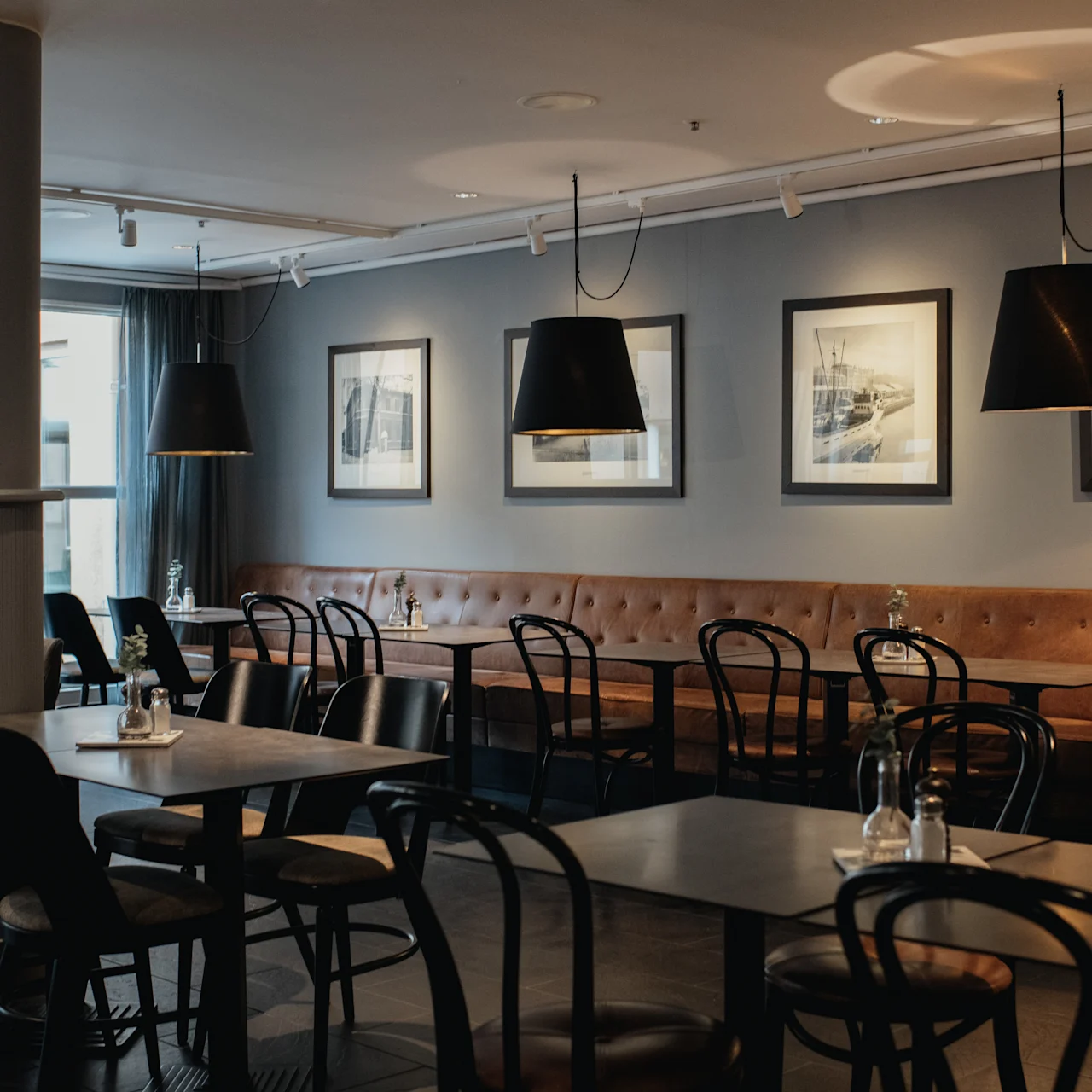 Tables with wooden chairs are set in a softly lit restaurant. Framed artwork hangs on light gray walls, and pendant lights illuminate the seating area. Large windows offer natural light.