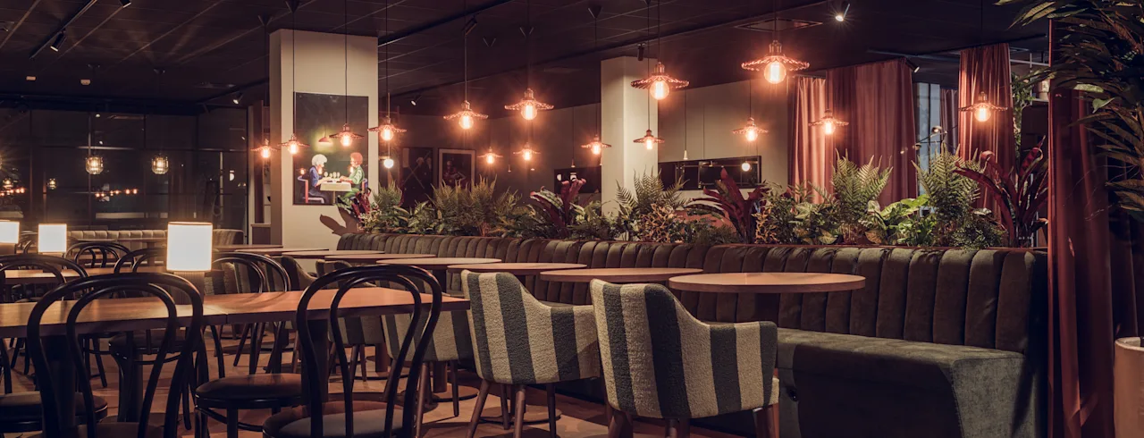 En restaurang har träbord och stolar arrangerade snyggt, omgiven av mjuk belysning. Krukväxter pryder utrymmet och skapar en mysig stämning med varma toner och modern inredning.