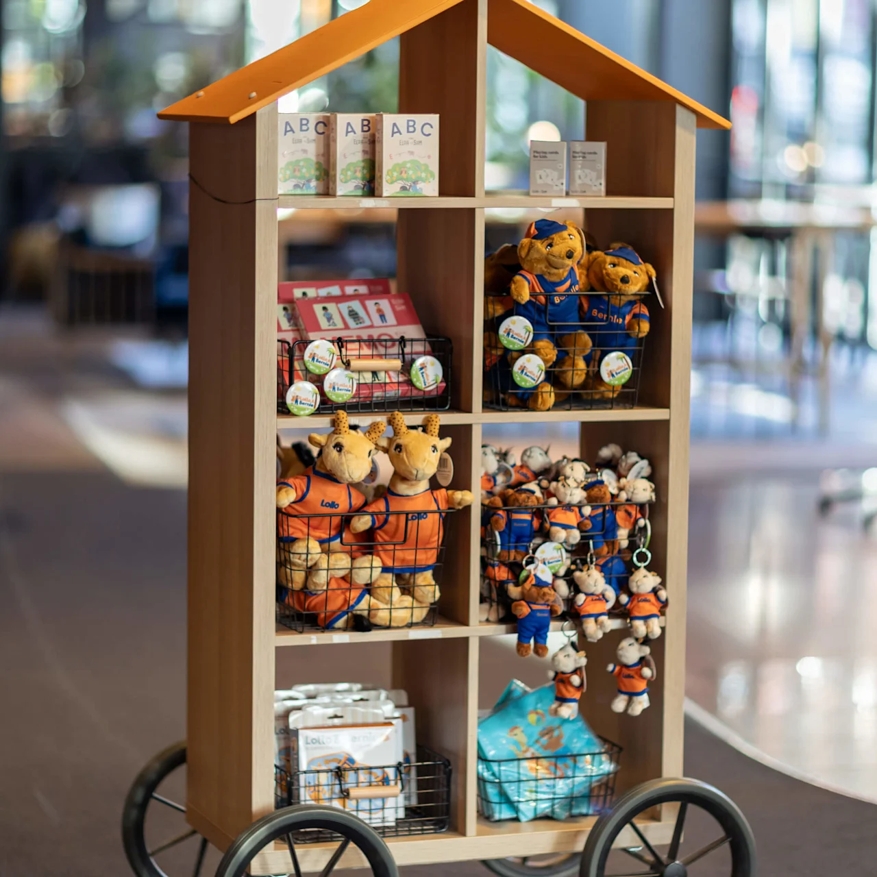 A wheeled display rack showcasing plush toys and books, including "ABC Lille Zoo." Situated indoors with a blurred background.