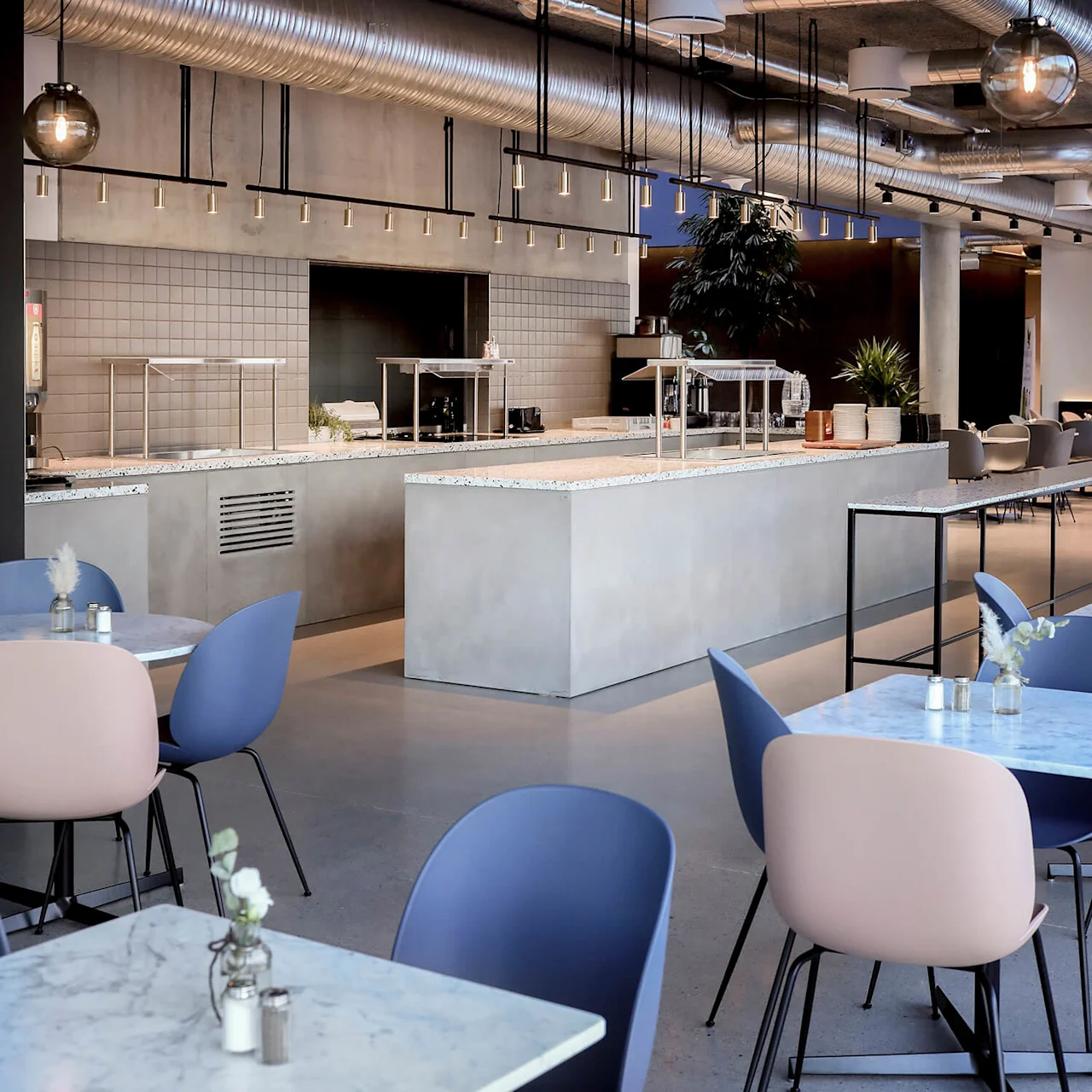 Moderne cafeteria med marmorborde og blandede pastelfarvede stole, med synlige metalliske loftsrør og blød belysning; planter og minimalistisk dekor komplementerer det åbne, industrielle miljø.