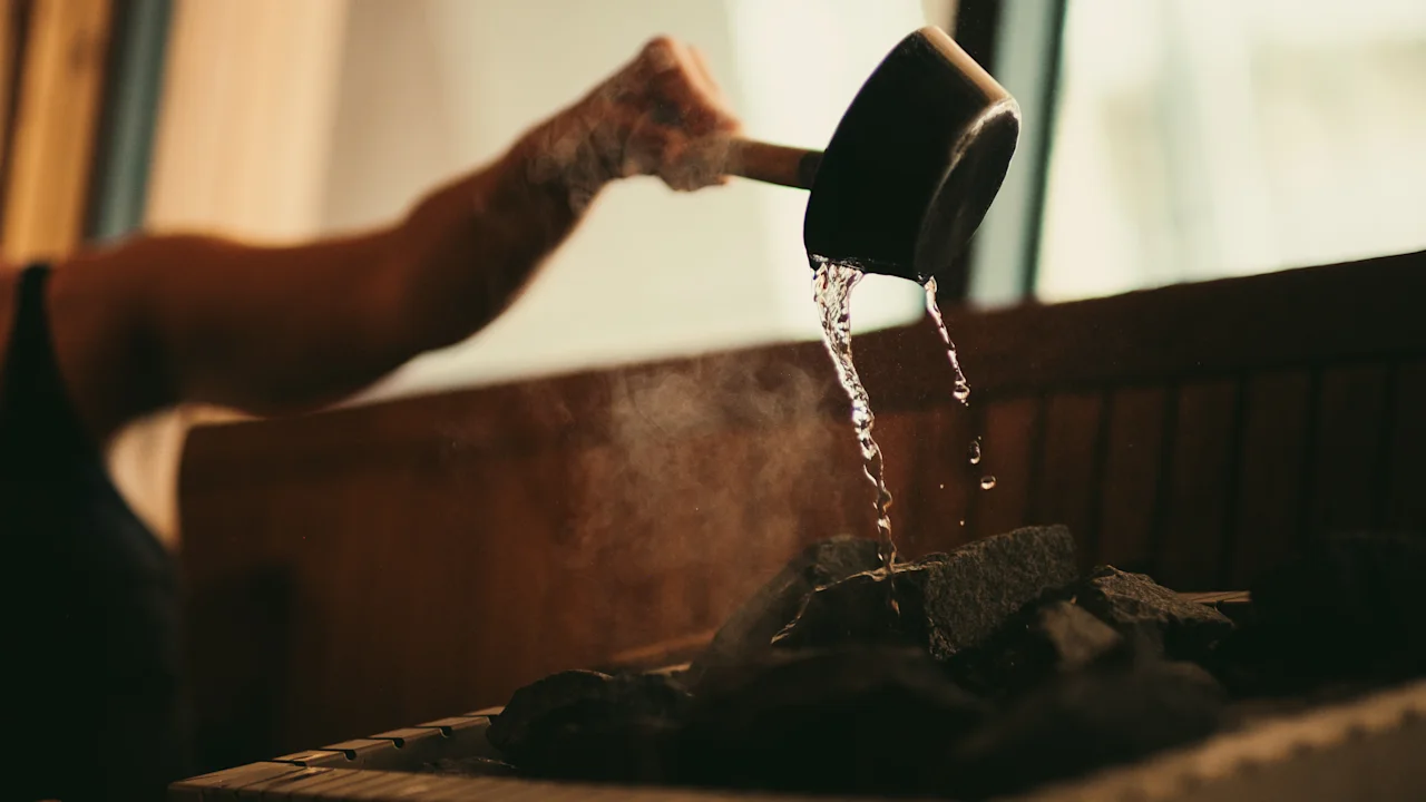 En hånd hælder vand fra en øse over varme sauna sten, hvilket skaber damp. Miljøet er en træsauna med naturligt lys gennem vinduer.