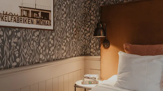Viihtyisä makuuhuoneen nurkkaus sängyllä, yöpöydällä ja kehystetyllä kuvalla kuvioidulla seinällä. Teksti: YKELFABRIKEN MONAR.
