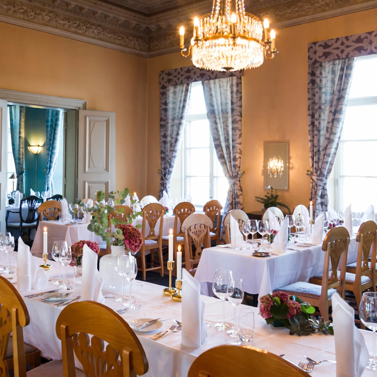 A dining room features elegantly set tables with white tablecloths, folded napkins, wine glasses, and floral arrangements. A chandelier hangs above, surrounded by large windows with patterned curtains.