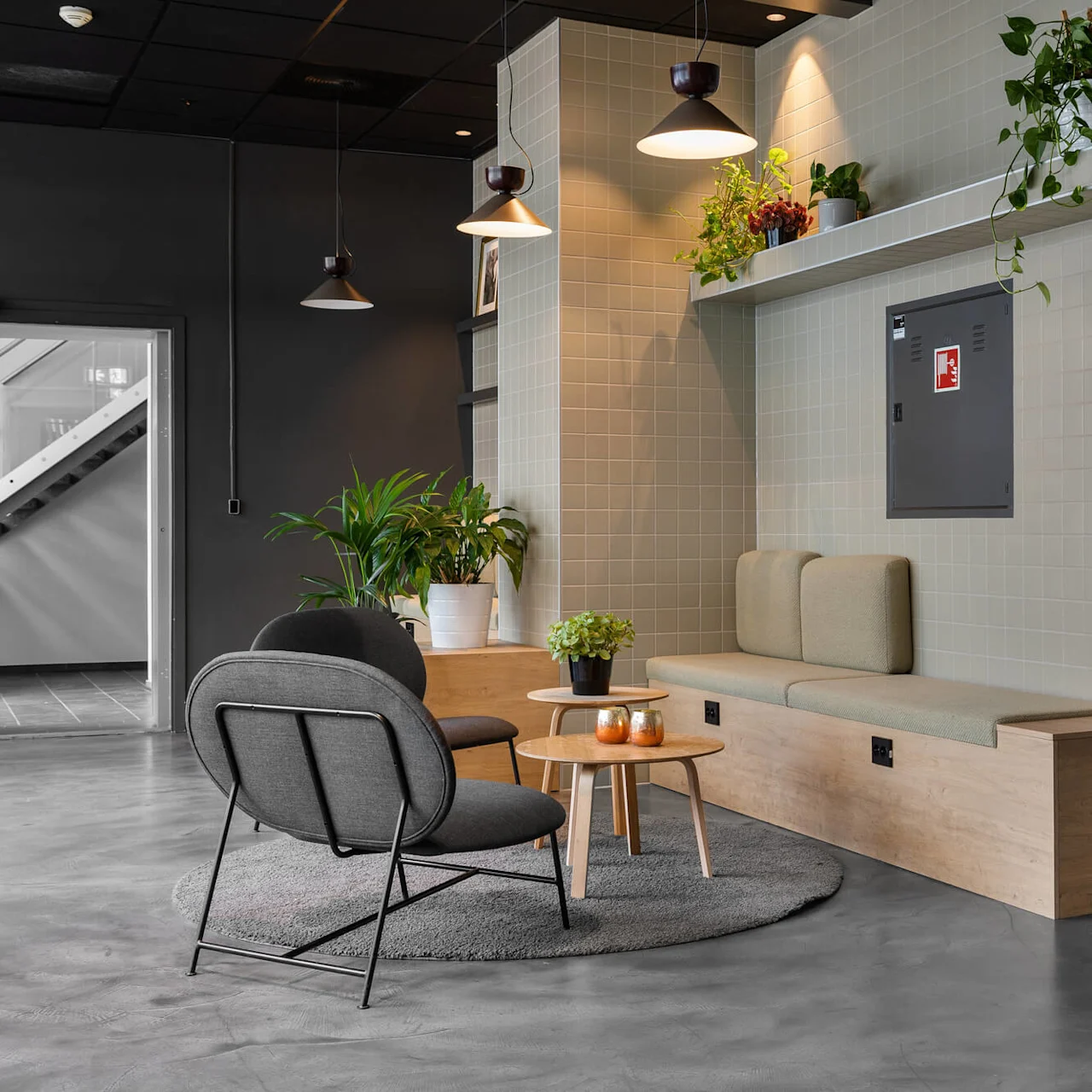 Cushioned bench with two pillows against a tiled wall featuring potted plants above. Adjacent, two gray chairs surround wooden tables. Nearby, a staircase and hanging lights. 