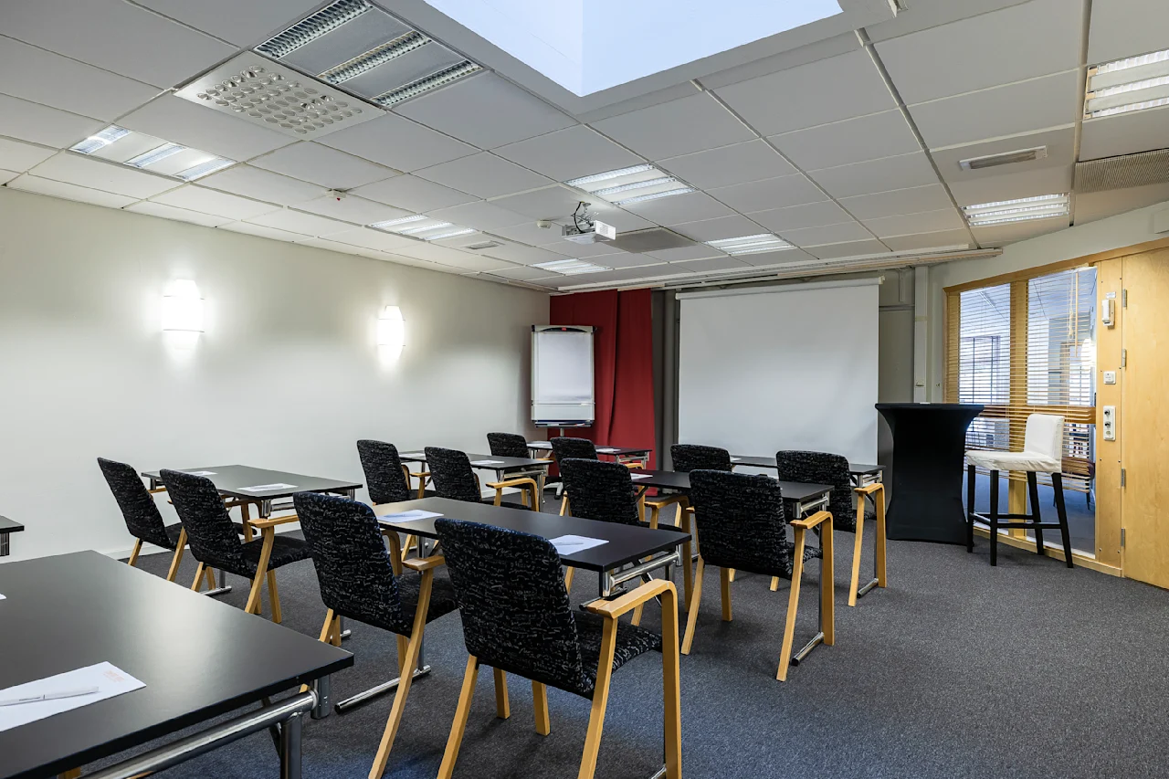 Et godt opplyst konferanserom med rader av tomme skrivebord og stoler vendt mot en hvit skjerm, klar for en presentasjon. En flippover står i nærheten, og et høyt bord er plassert på siden.