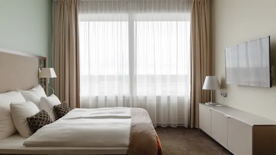 Ett mysigt hotellrum med en bekväm säng, ett stort fönster med gardiner och en väggmonterad TV.