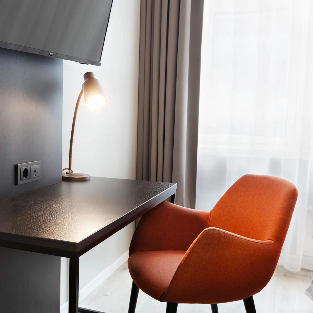 Et træbord med en tændt lampe, orange stol og vægmonteret TV i nærheden, placeret ved et vindue med lyse gardiner. To klare glas står på bordet.