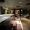 A luxurious indoor spa area with a large bubbling hot tub, lounge chairs, and a serene swimming pool in the background.