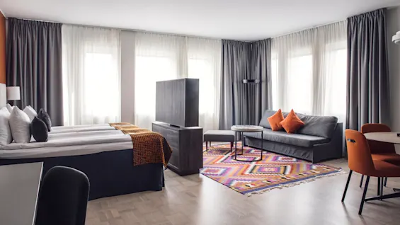 Ett modernt hotellrum har en dubbelsäng och soffa, dekorerade med orange accenter. En färgglad geometrisk matta och fönster från golv till tak skapar en ljus, inbjudande atmosfär.