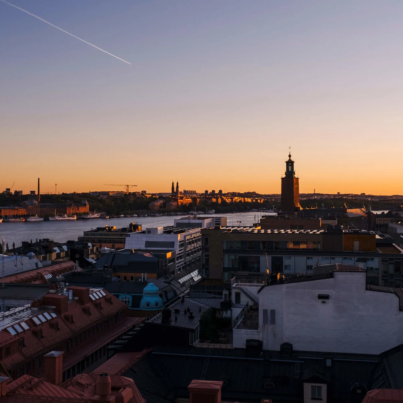 Skyline at sunset features a clock tower silhouetted against an orange sky, above a cityscape of rooftops and buildings, next to a calm river under a clear sky with a contrail.