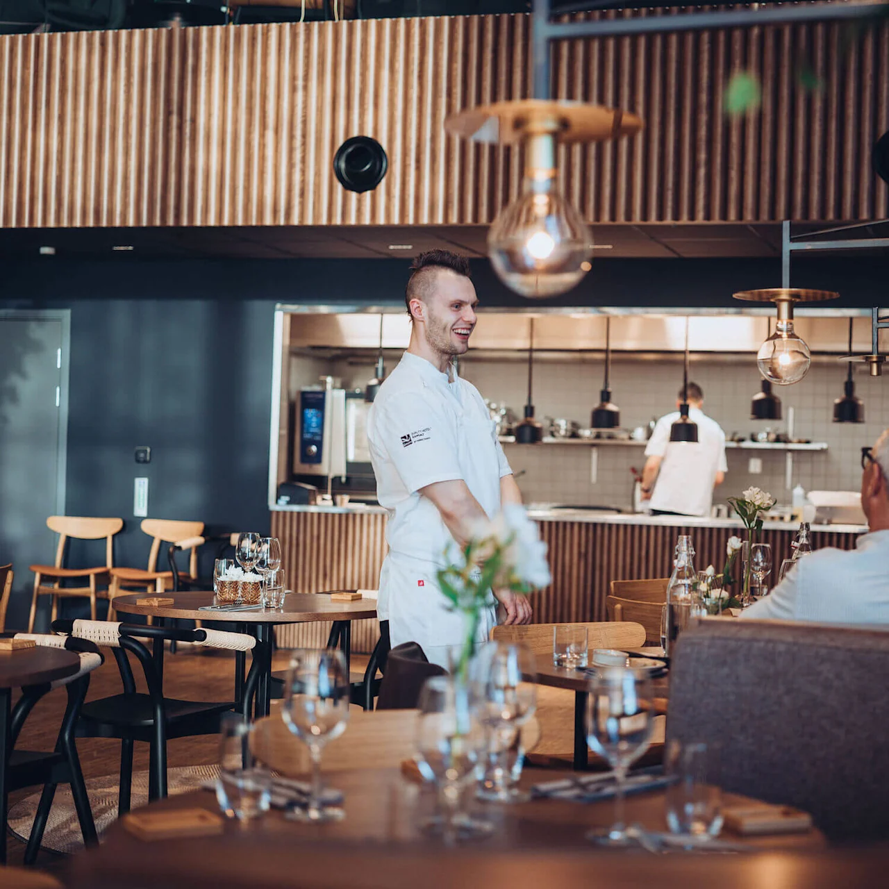 A smiling chef stands in a modern restaurant, engaging with diners seated at wooden tables. Contemporary lighting and a visible kitchen in the background contribute to the warm atmosphere.