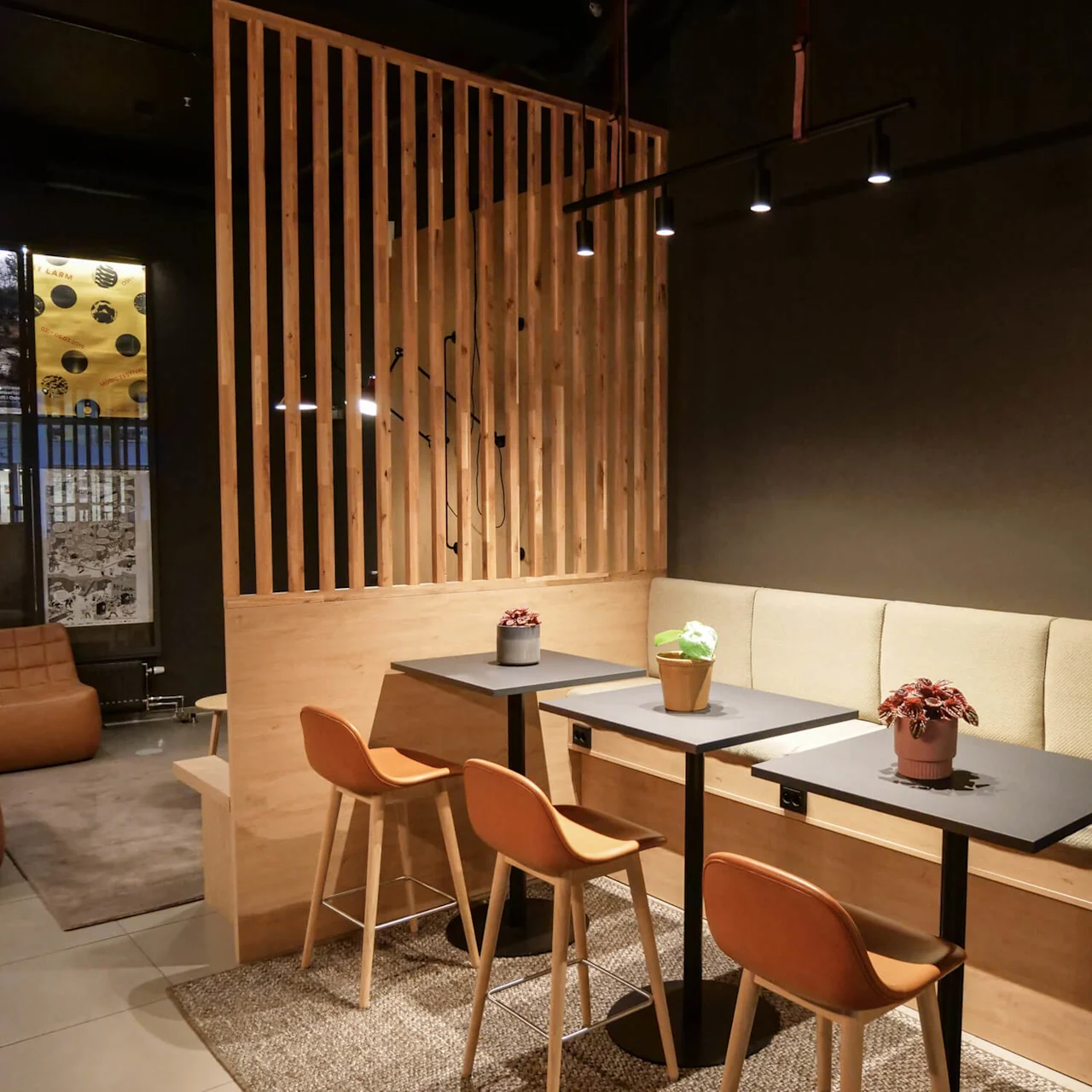 Two small tables with orange chairs and potted plants sit in a cozy seating area, featuring a soft bench against a dark wall, lit by modern overhead lights.