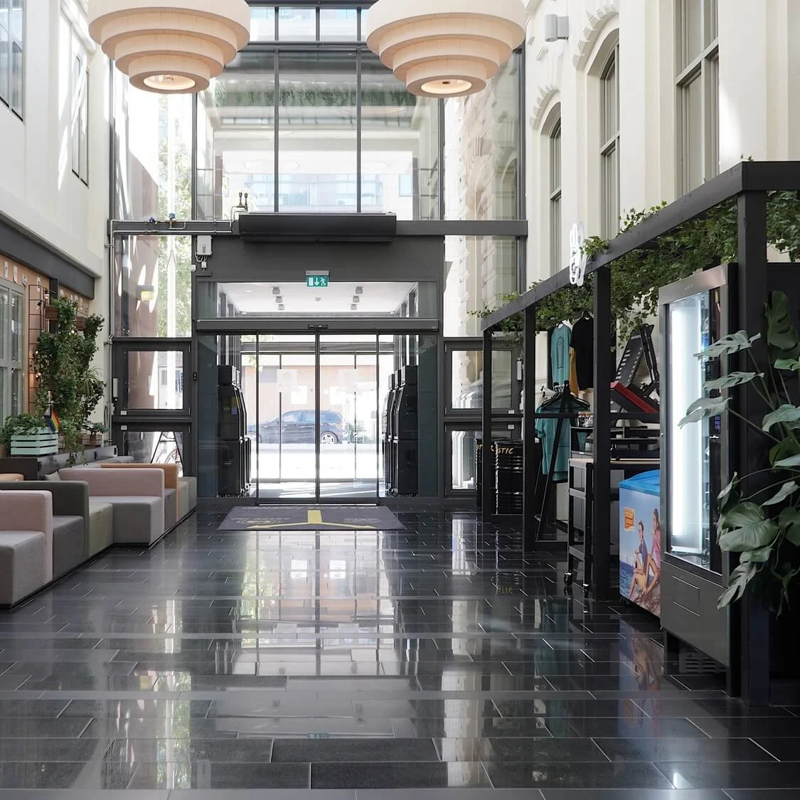 An indoor lobby with sofas, potted plants, and a glass entrance door, reflecting a glossy tiled floor.