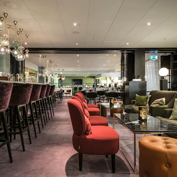 Luksusbar med lilla barstolar och ljus på vänster sida, medan röda stolar och plyschsoffor skapar en mysig sittgrupp på höger sida, under eleganta hänglampor.
