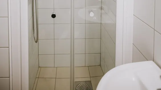 Brusekabine med regnbruser og sæbedispenser, omgivet af hvide flisevægge og en glasdør i et badeværelsesmiljø.