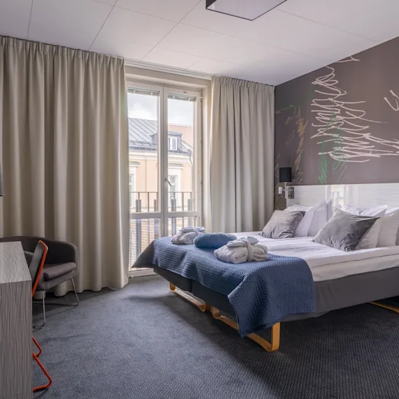 Ett hotellrum har en snyggt bäddad dubbelsäng med blå och grå sängkläder, prydd med två vikta handdukar. Gardiner täcker ett fönster och kompletterar den moderna inredningen.